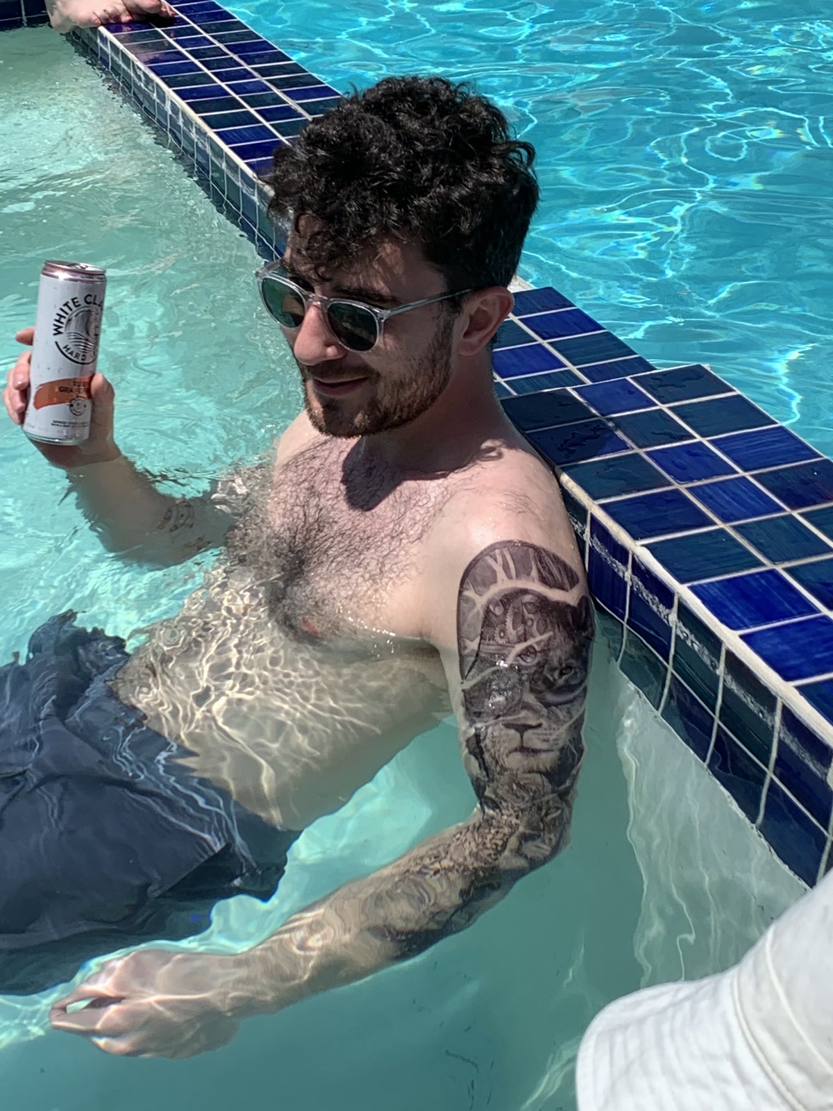 Alex in a pool, sunglasses on, full sleeve tattoo out, White Claw in hand